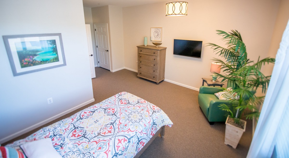 A cozy bedroom featuring a patterned bedspread, wooden dresser, wall-mounted TV, and a green armchair.
