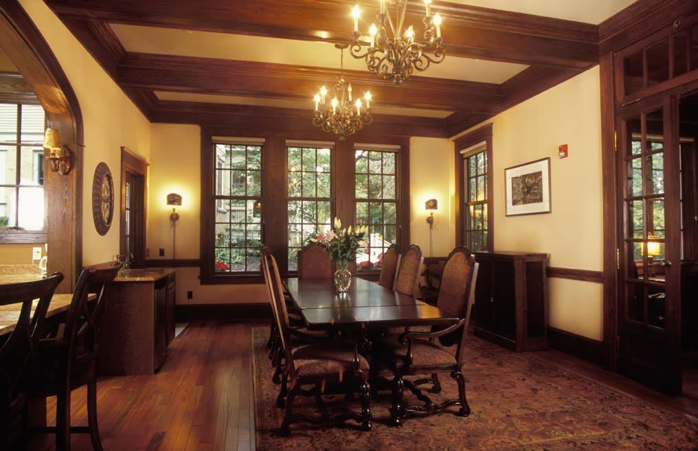 A traditional dining room with wooden furniture and chandeliers, featuring large windows and a floor rug.