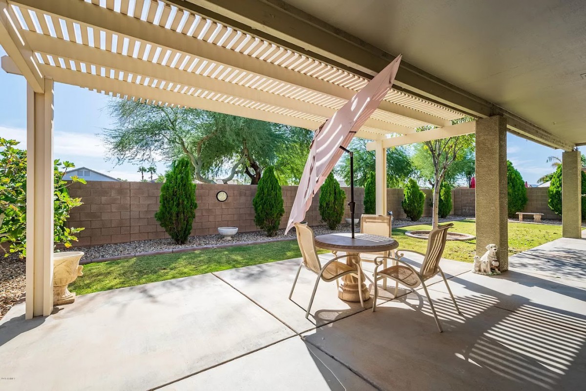 A sunny backyard patio with a table, chairs, and an umbrella set on a concrete floor surrounded by a lush garden.