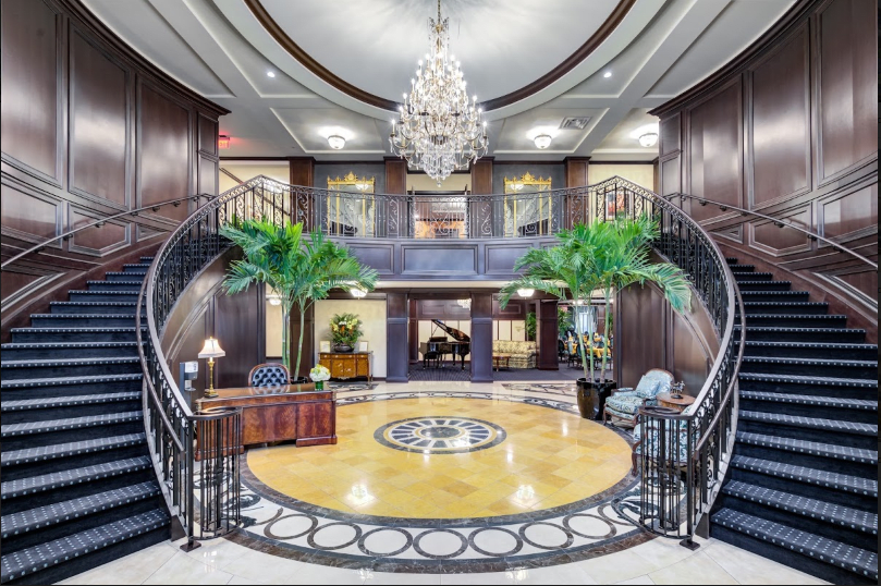 A luxurious hotel lobby featuring a grand staircase, elegant chandelier, and lush palm plants.