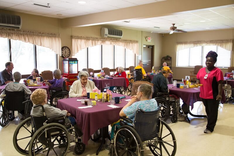 Elderly residents gather in a nursing home dining area, seated around tables, enjoying a communal meal.