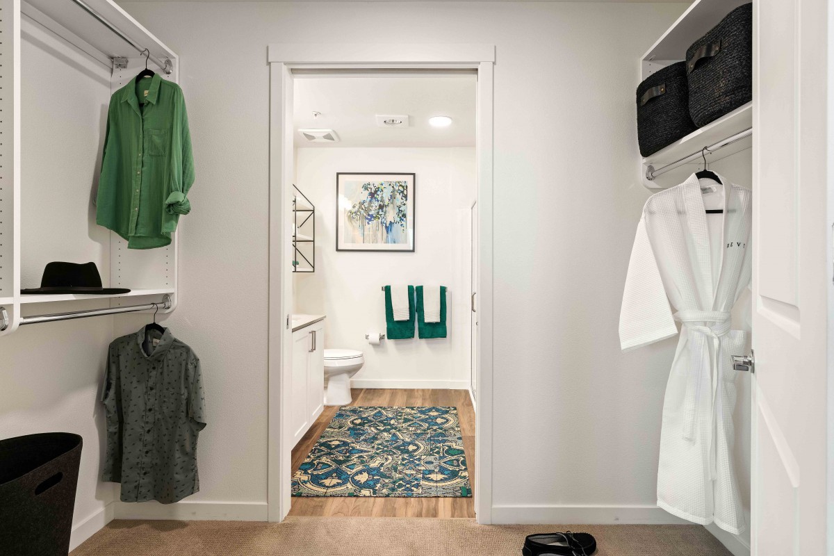 A view from a walk-in closet into a bathroom, with clothes hanging on either side and a decorative rug leading to the bathroom.