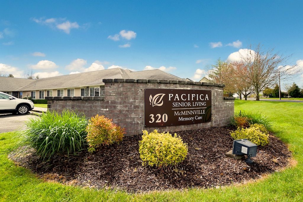 A view of a sign for Pacifica Senior Living with landscaping and a building in the background.