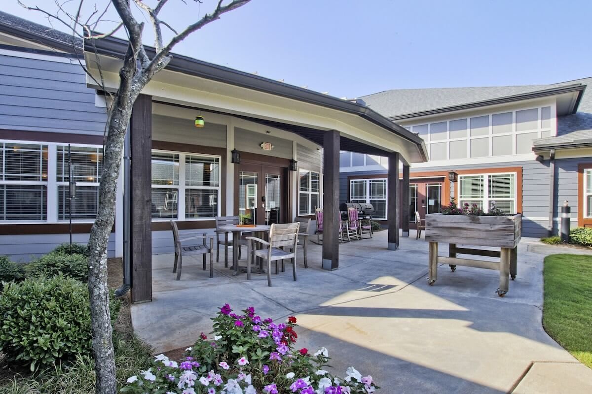 A cozy outdoor patio area with tables, chairs, and colorful flowers, adjacent to a modern building.