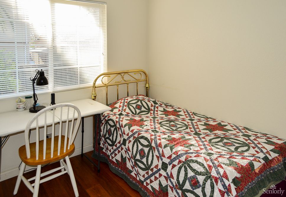 A cozy bedroom with a single bed adorned with a patterned quilt, next to a small desk and chair by the window.