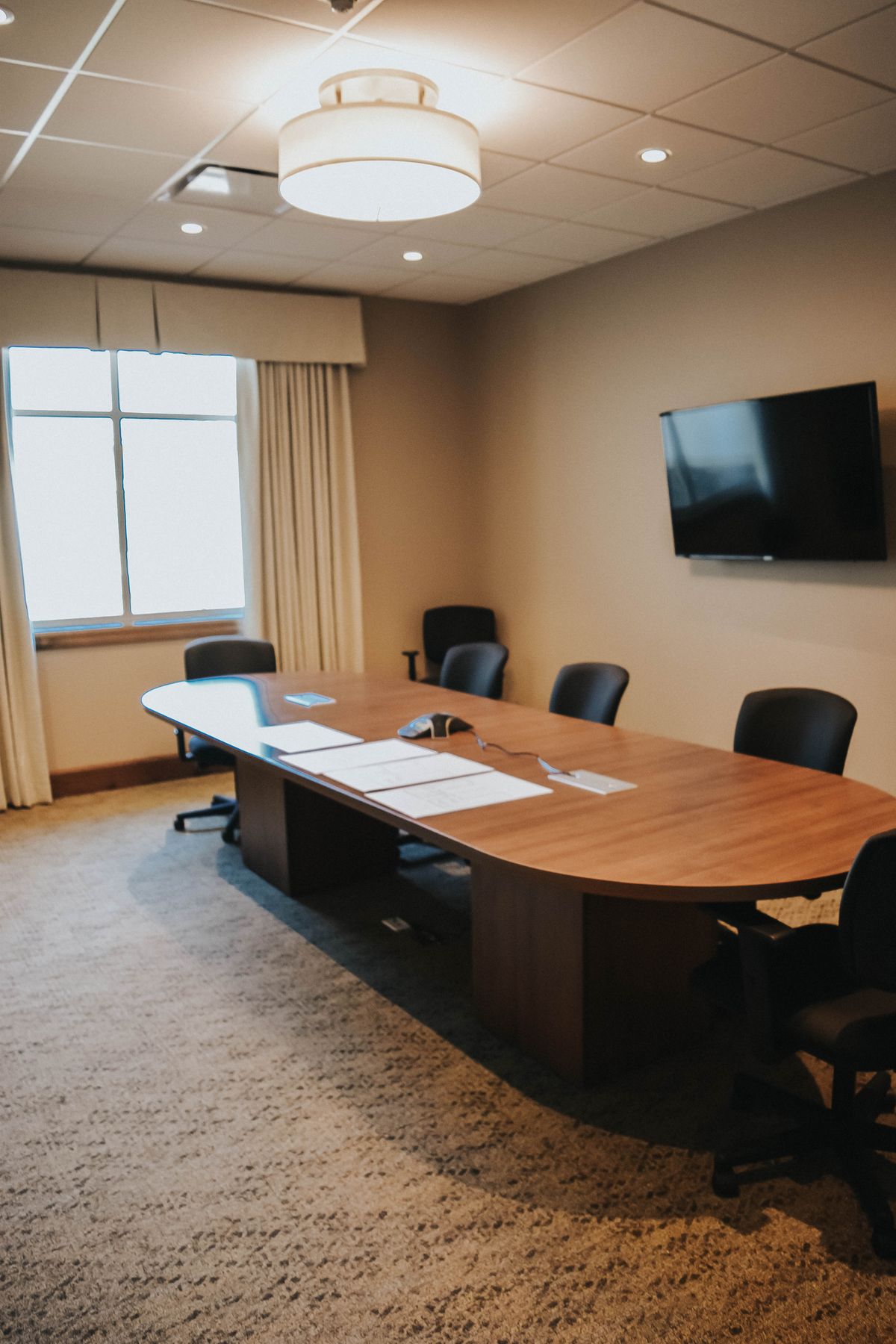 A modern conference room with a wooden table, several chairs, and a wall-mounted television.