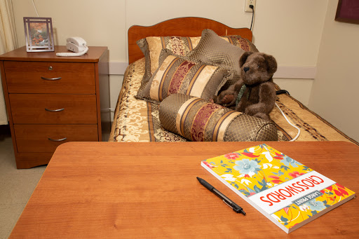 A cozy bedroom scene featuring a neatly made bed with decorative pillows, a stuffed animal lounging on the bed, and a study desk holding a colorful children's book.