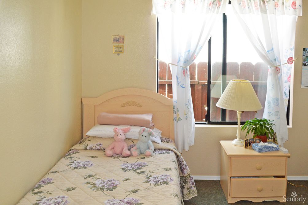 A cozy bedroom with a single bed, adorned with stuffed animals and floral bedding, next to a window letting in sunlight.