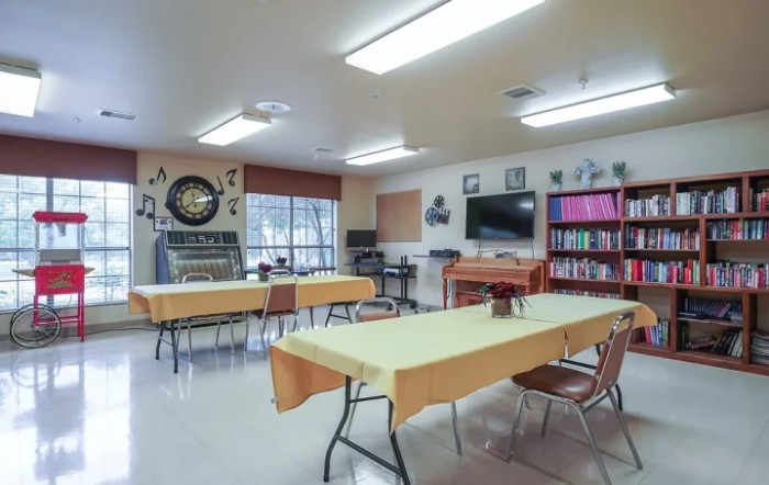 A classroom setup with tables and chairs, featuring a whiteboard, bookshelves, and wall decorations.