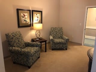 A cozy living room corner featuring two patterned armchairs and a small table with a lamp, accompanied by framed artwork on the wall.