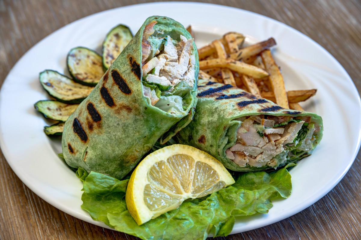 A grilled chicken wrap served with zucchini, fries, and a lemon wedge on a bed of lettuce.