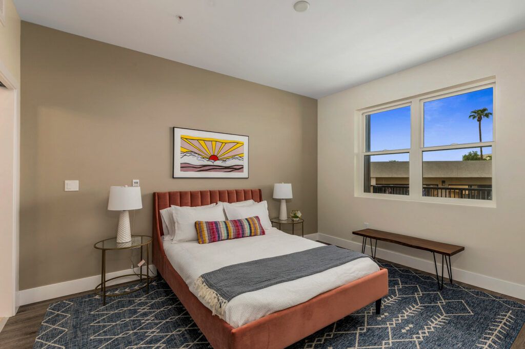 A cozy bedroom featuring a bed with colorful pillows, two bedside lamps, a window, and wall artwork.