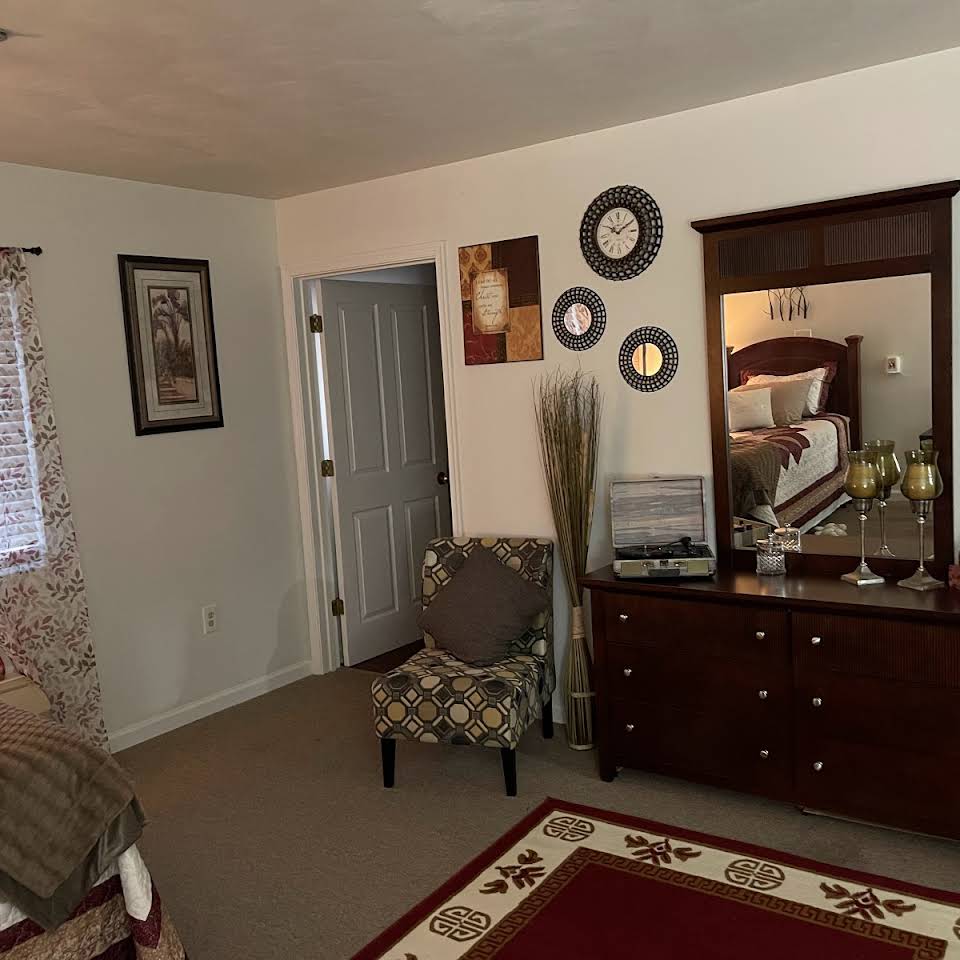 A cozy bedroom featuring a dresser with a mirror, adorned with decorative items, and a patterned chair next to the wall artwork.