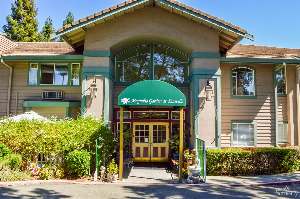 The entrance of a building with a green awning, surrounded by greenery on a sunny day.