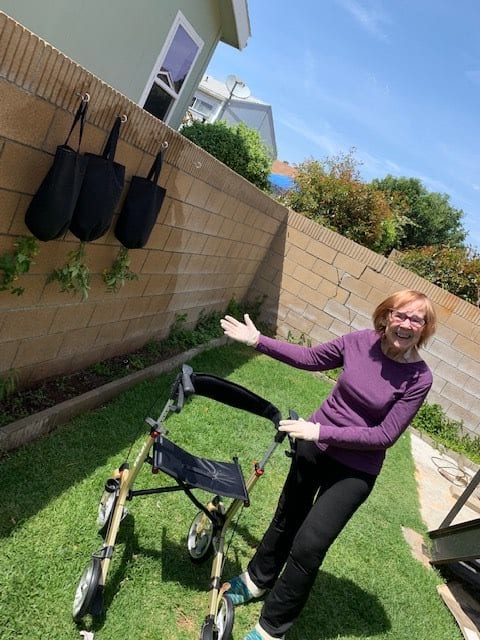 A woman smiling in a garden with a walker and plants hanging on a wooden fence.