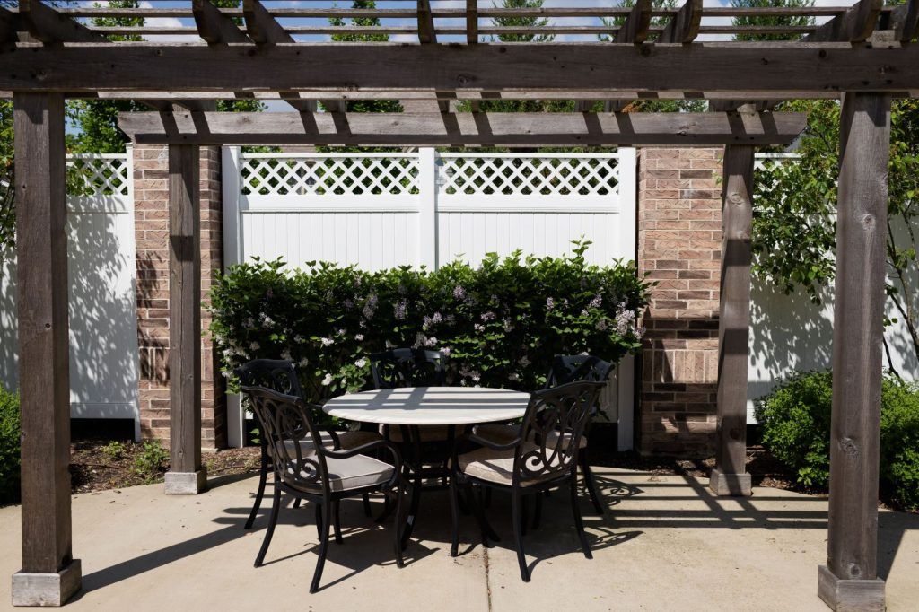 A patio area with a round table and four chairs under a pergola, surrounded by greenery.