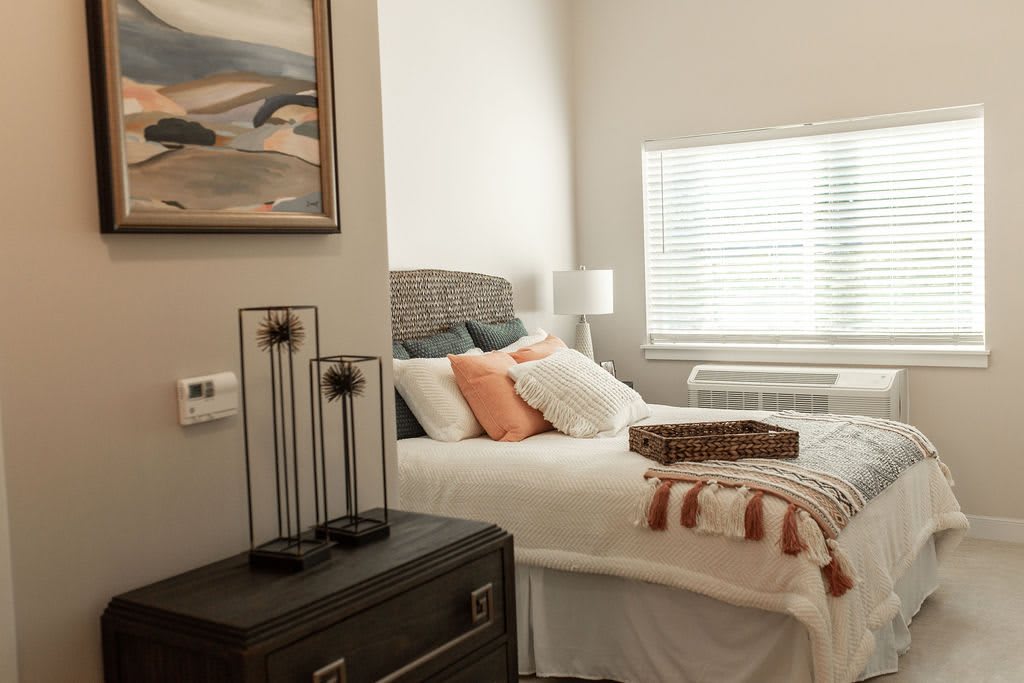 A cozy bedroom with decorative pillows and a soft blanket, featuring a bedside lamp and wall art.