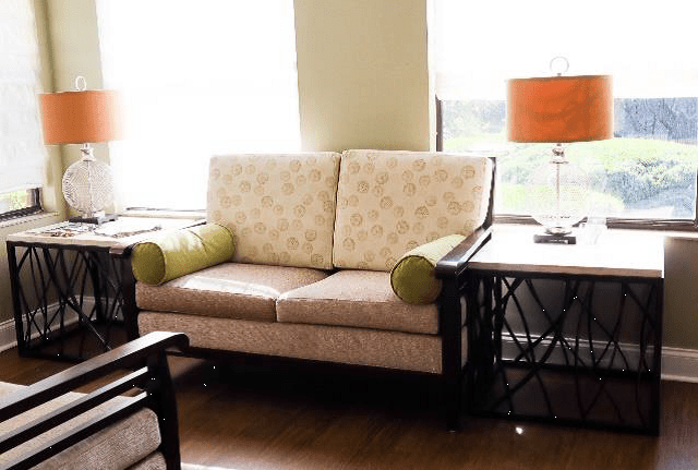 A cozy living room setup featuring a patterned sofa, side tables, and a bright lamp near large windows.