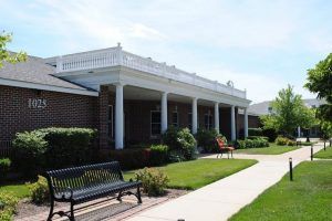 A view of a building with columns and surrounding landscaping, including a bench and sidewalk.