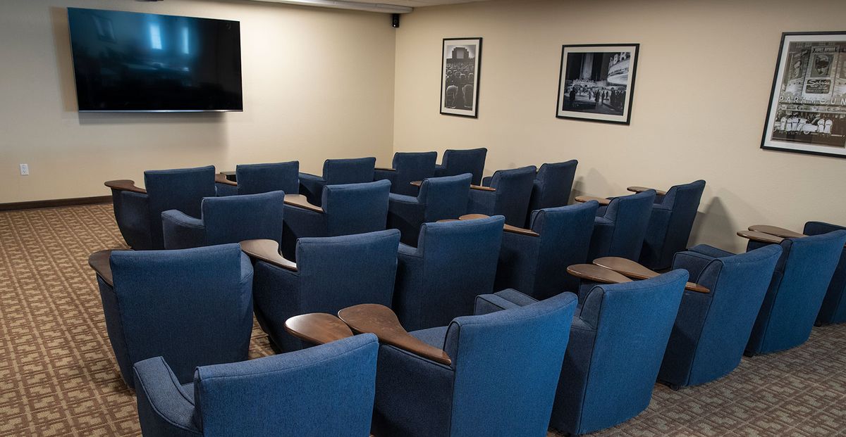A small theater room with blue upholstered chairs, each equipped with a wooden table, facing a television screen and decorated with framed wall art.