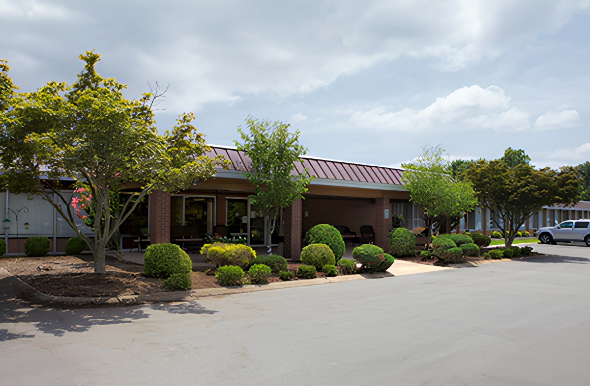 A single-story brick building surrounded by well-maintained landscaping, with a parking area in the foreground and a cloudy sky above.