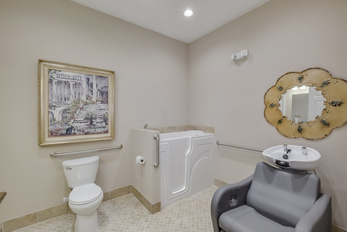 A bathroom featuring a walk-in tub, toilet, and salon-style chair with a decorative mirror and framed artwork on the wall.