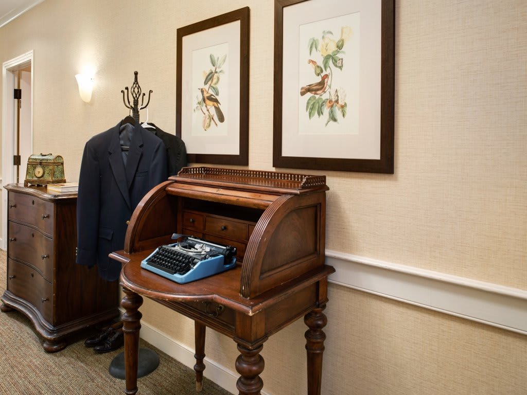 A vintage typewriter sits atop a wooden roll-top desk, with framed artwork and a suit hanging nearby.
