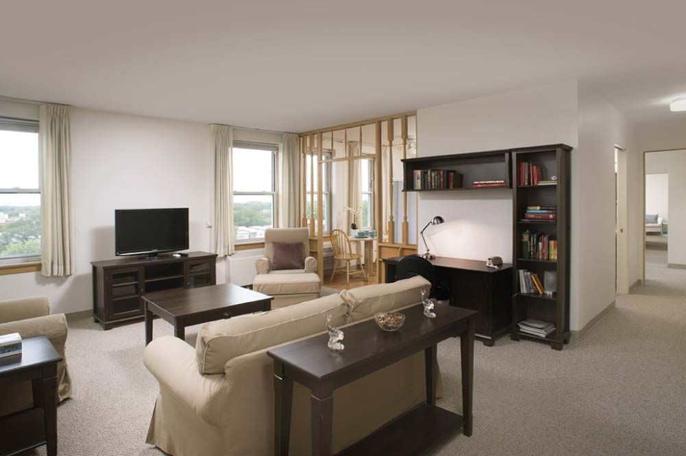 A cozy living room with a sofa, coffee table, and a television, featuring neutral colors and functional furniture such as a desk and bookshelf.