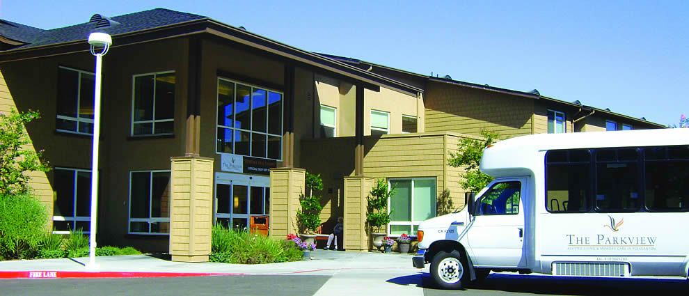A building entrance with a parked van and surrounding greenery on a sunny day.