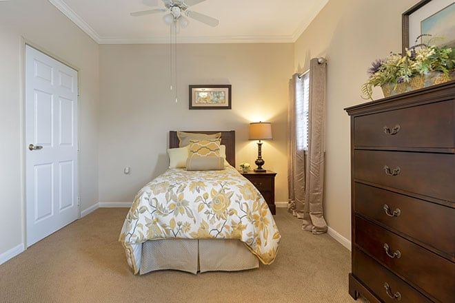 A cozy bedroom with a single bed, nightstand, and dresser, softly lit by a table lamp.