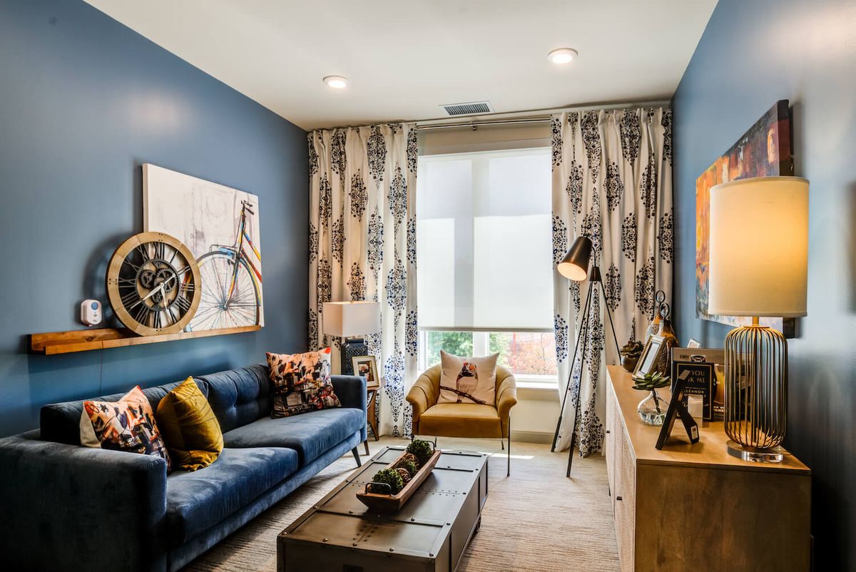 A modern living room with blue walls and stylish decor, featuring a sofa, artwork, and a large window with patterned curtains.