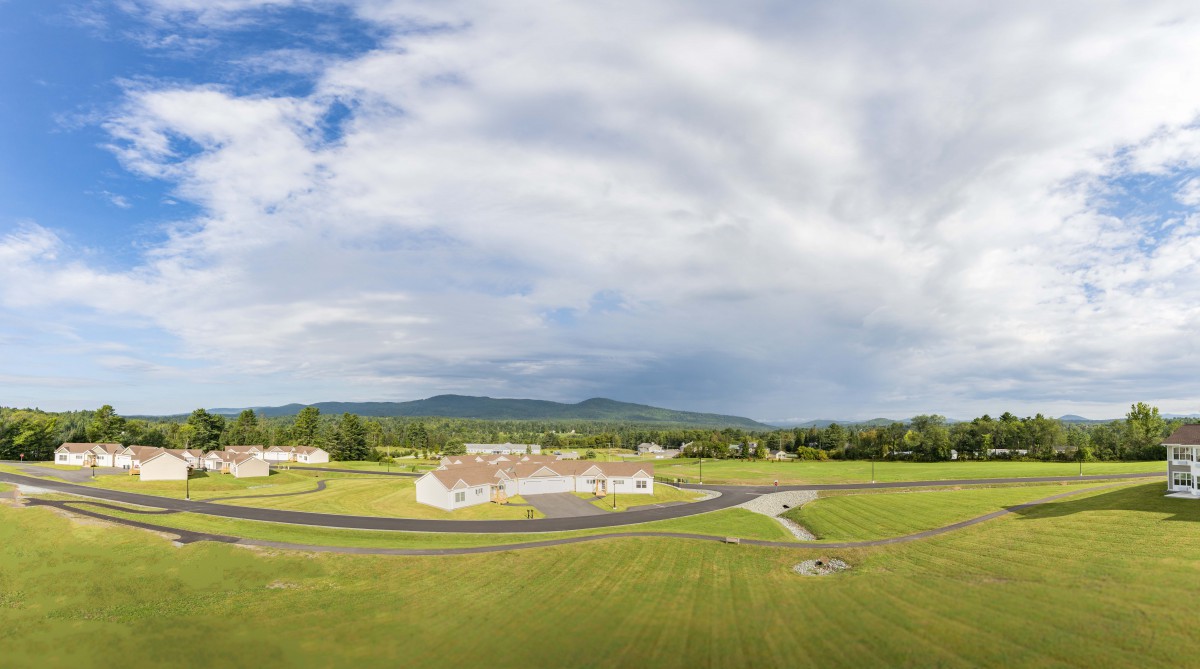 A scenic view of buildings surrounded by green fields under a cloudy sky, with mountains in the background.