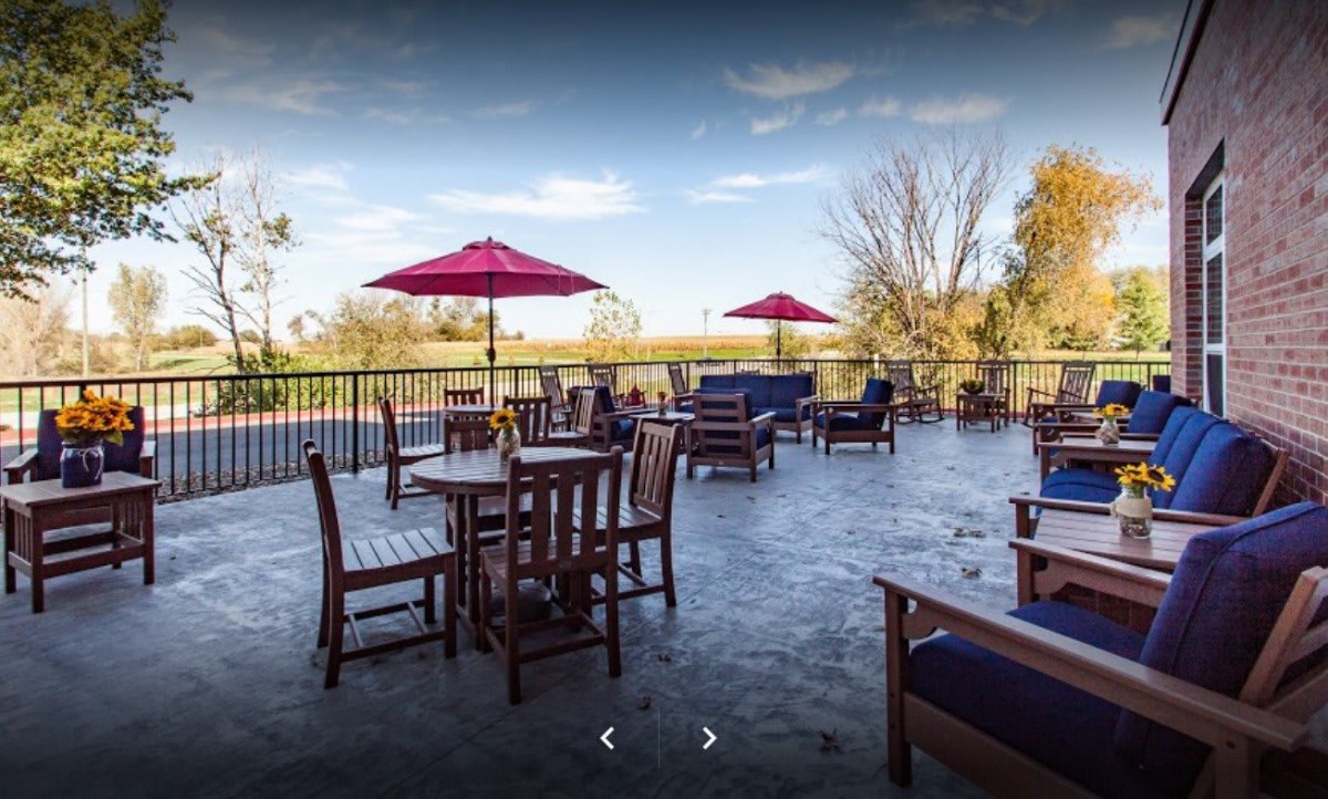 A spacious outdoor patio with wooden tables, chairs, and red umbrellas, set against a scenic backdrop.