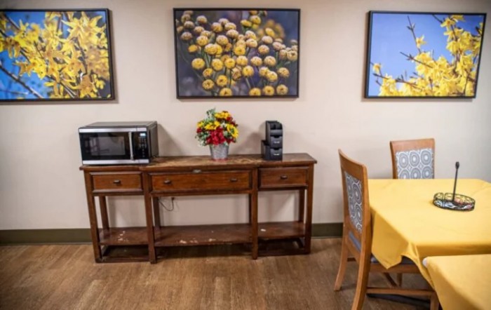 A cozy kitchen area with a wooden table, microwave, coffee machine, and decorative flower paintings on the wall.