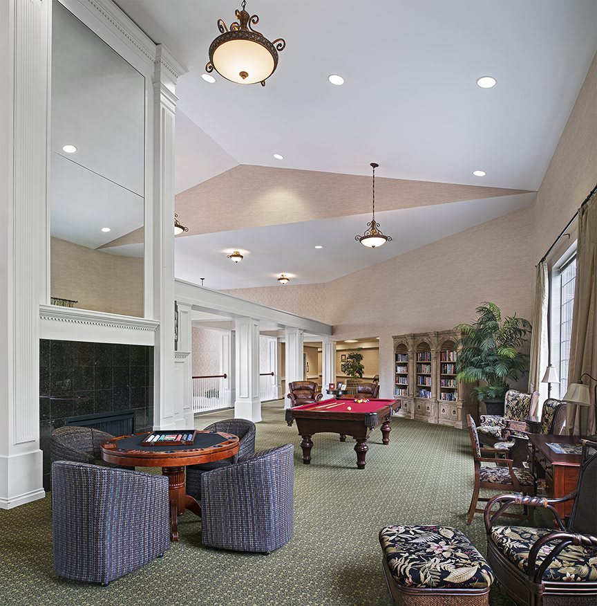 A spacious recreation room featuring a pool table, lounge chairs, and tables, with elegant lighting and large windows.
