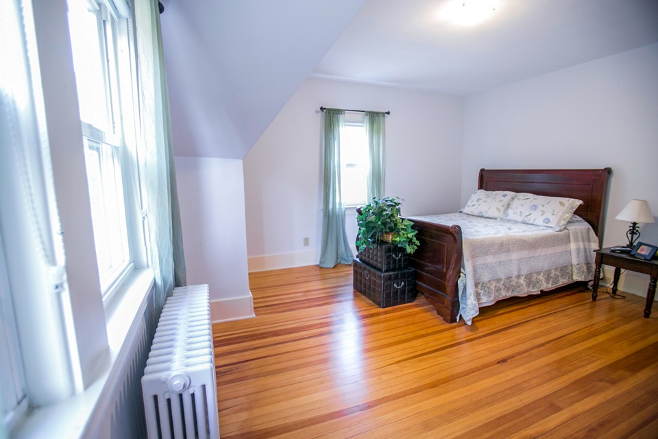 A cozy bedroom with hardwood floors, featuring a bed, indoor plant, and natural light from a window.