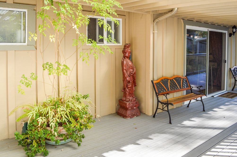 A serene outdoor patio with a decorative statue, wooden bench, potted plant, and wooden siding under a shaded area.