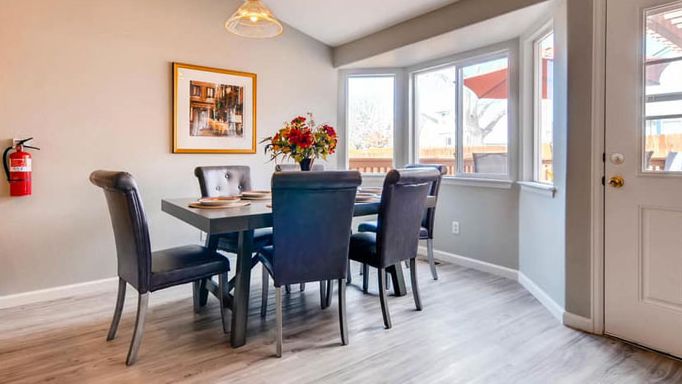 A modern dining room with a table and chairs set, adorned with a floral centerpiece near large windows.