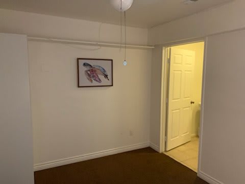 An empty room with a ceiling fan, framed artwork on the wall, and an open door leading to a lit hallway.