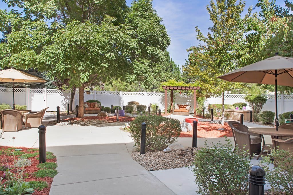 A tranquil garden setting with a paved pathway, seating area, lush trees, and a pergola.