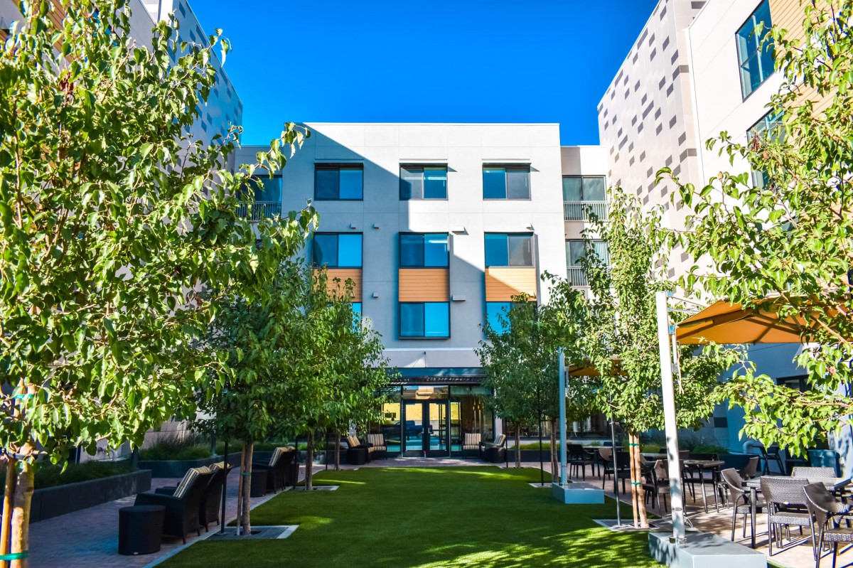 A modern apartment building surrounded by trees and a well-kept lawn under a clear blue sky.