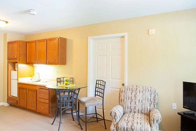 A cozy interior space featuring a small kitchenette with wooden cabinets, a high table with chairs, and a comfortable armchair.