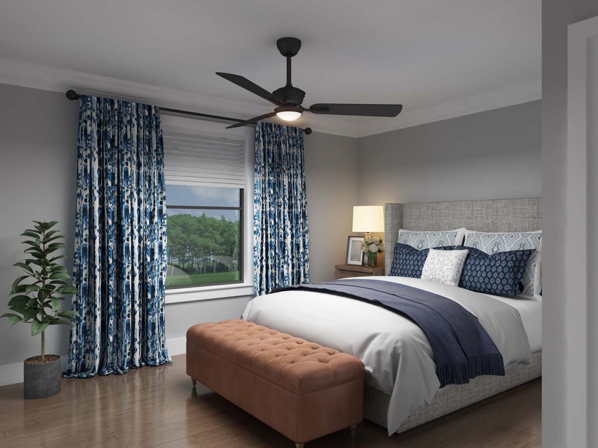 A cozy and elegantly furnished bedroom featuring a ceiling fan, large window with blue patterned curtains, a well-made bed with blue accents, and a bedside lamp.