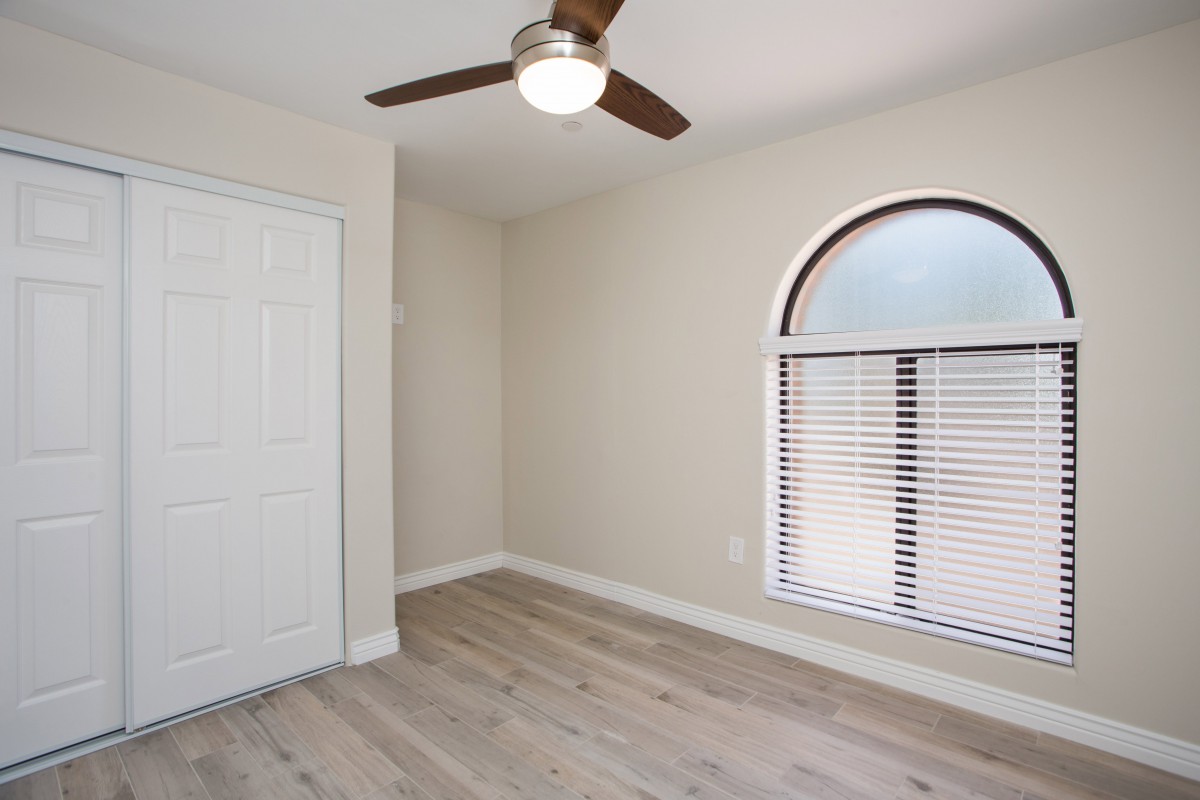 A small empty room with a ceiling fan, wood flooring, and an arched window with blinds.