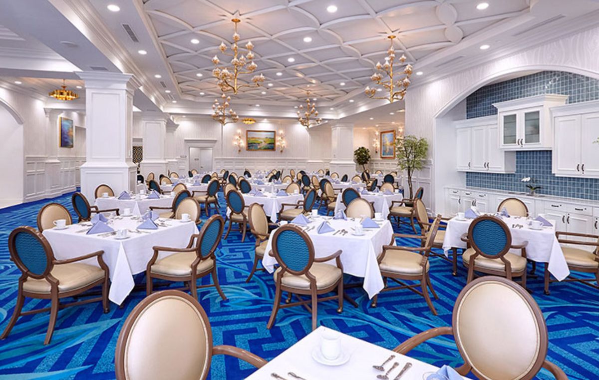 An elegant restaurant dining room with blue carpeting and white tablecloths, featuring classic seating and chandeliers.