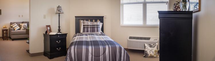 A neatly arranged bedroom with a single bed featuring striped bedding, next to a window and an air conditioner.