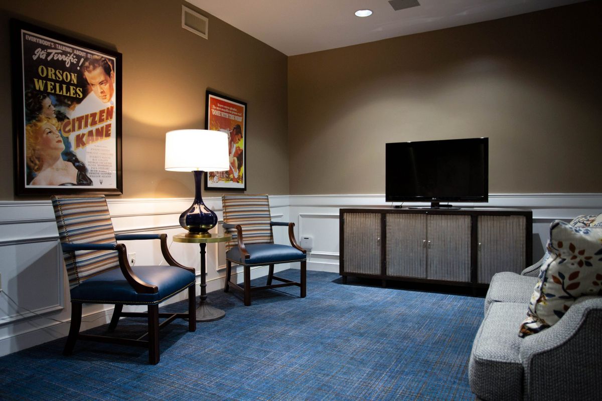 A cozy living room with a television, chairs, and a table lamp on blue carpet flooring.