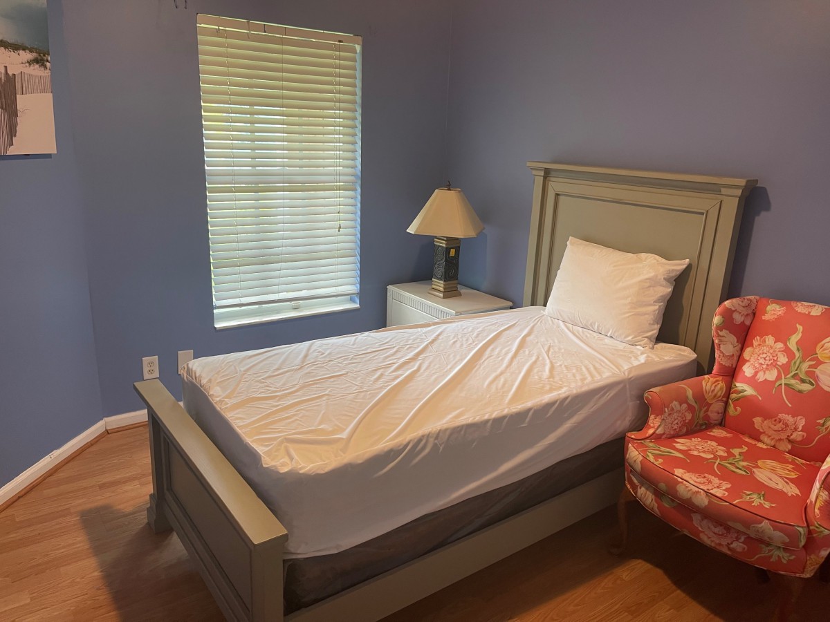 A small, cozy bedroom with a single bed, nightstand, lamp, and chair next to a window with blinds.
