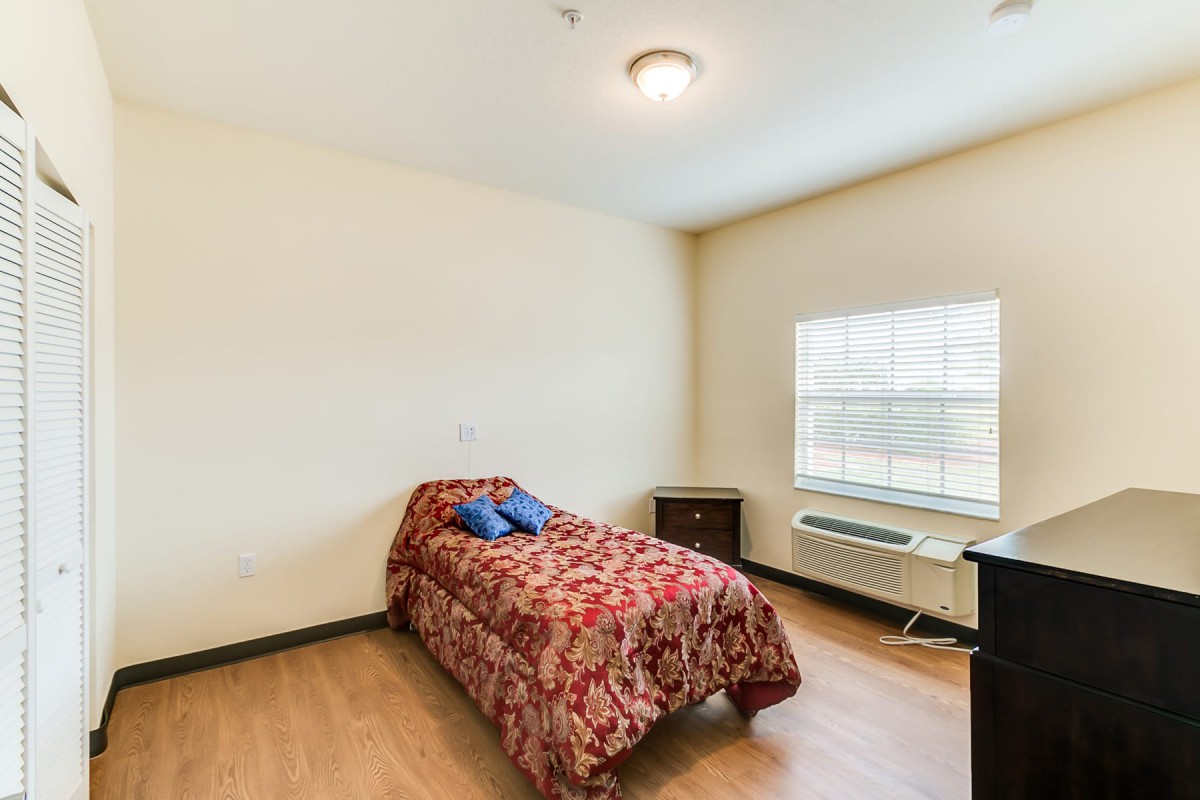 A simple bedroom featuring a single bed with a floral cover, a bedside table, and a window letting in natural light.