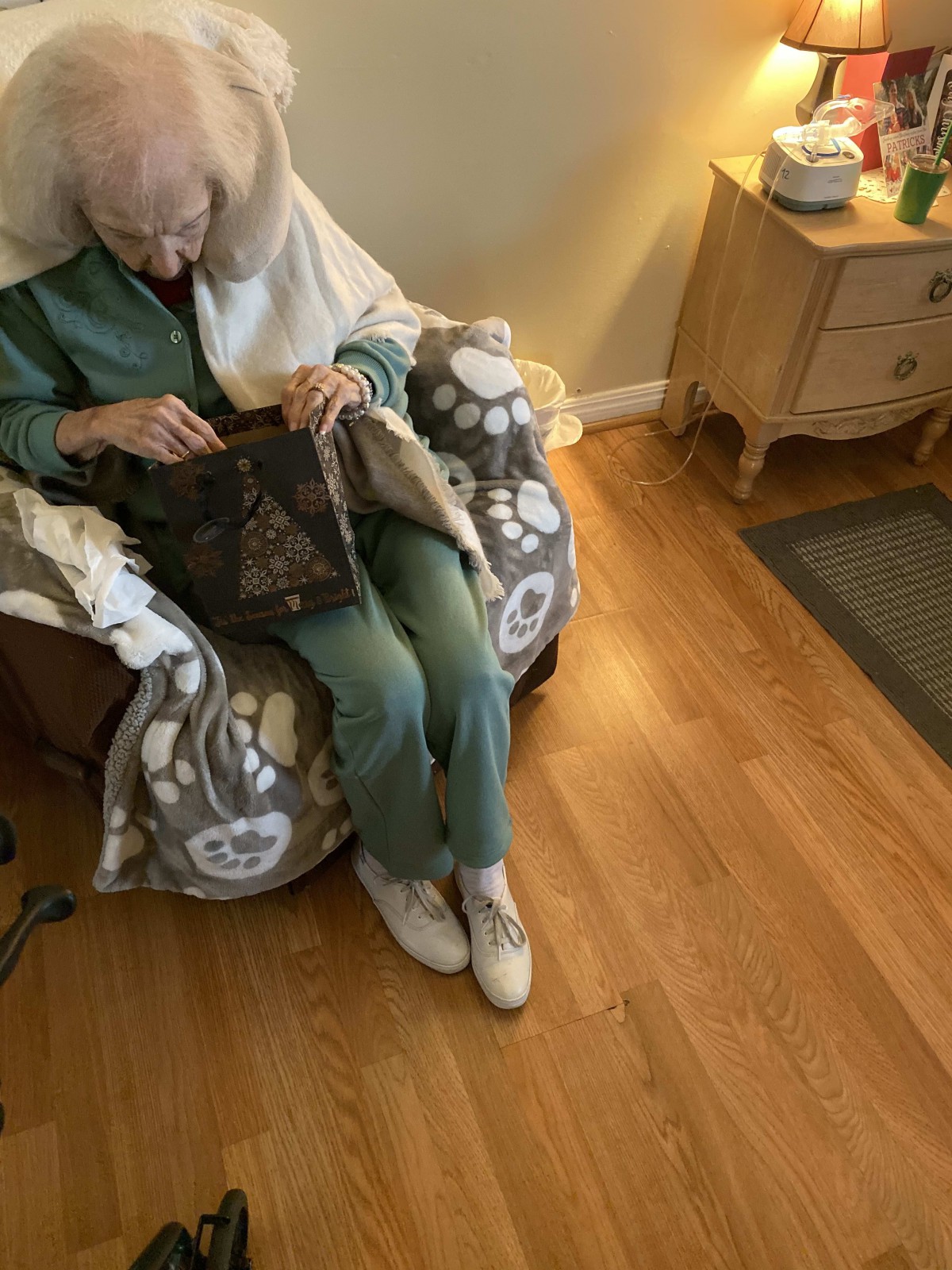 An elderly person sits in a cozy chair, knitting under a blanket in a warmly lit room.